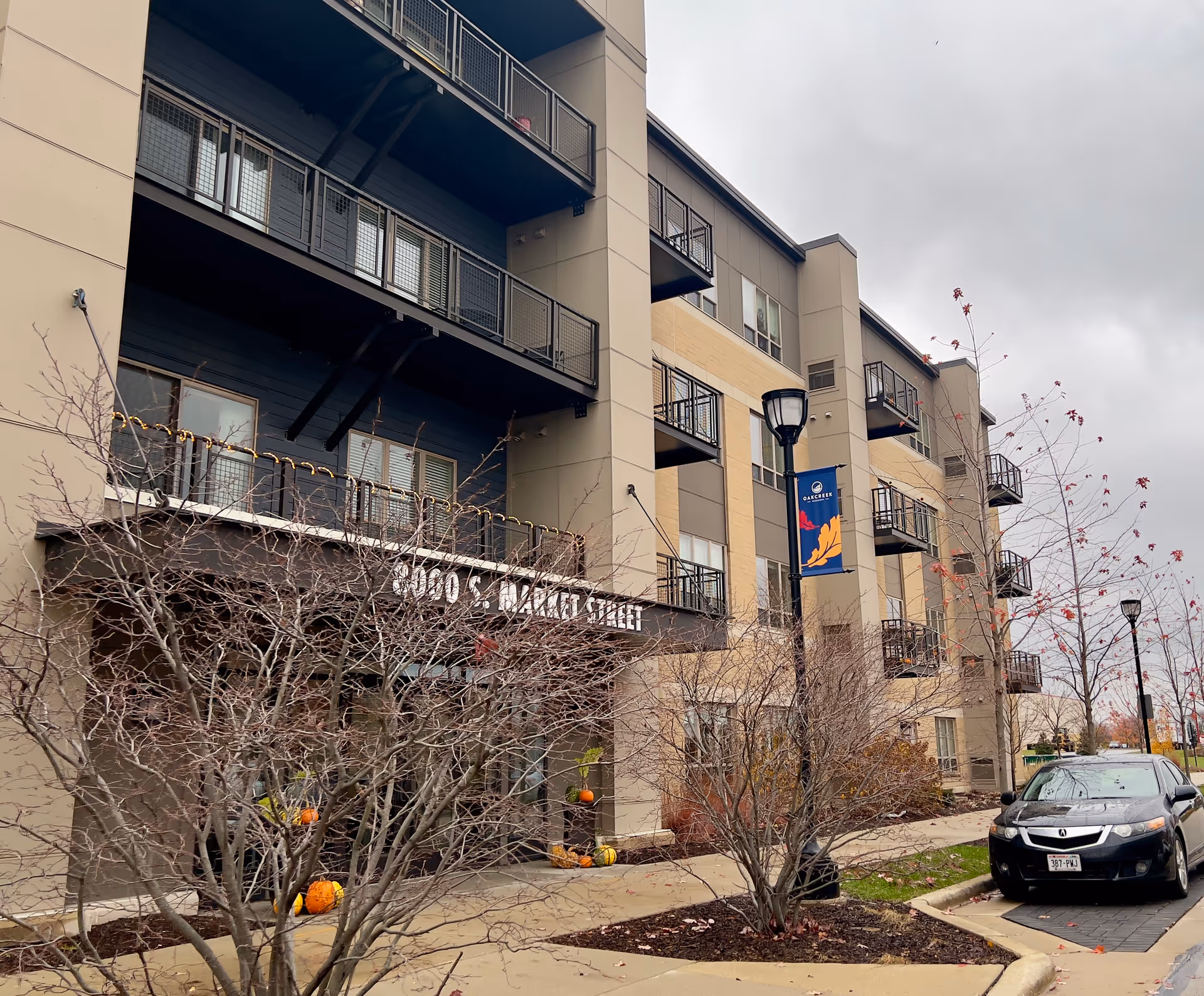 Exterior view of a multi-story residential building with balconies, leafless trees, a sidewalk, a street lamp with a banner that reads 'Oak Creek', and a parked black car under a cloudy sky.