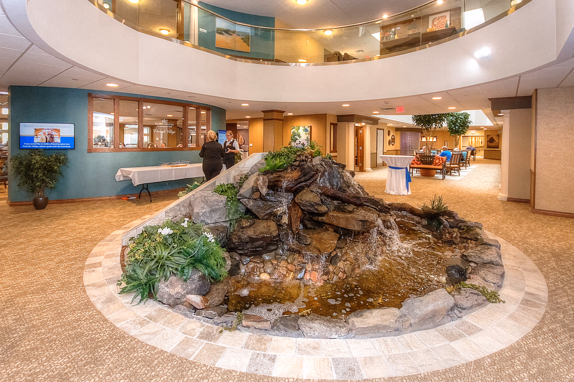 Indoor common area of Parkside Village Retirement Resort featuring a large decorative rock waterfall with plants in the center. Two people are conversing near a windowed office area with a blue wall and a TV screen. The space has carpeted floors, seating areas with chairs and tables, and a second-floor balcony overlooking the area.