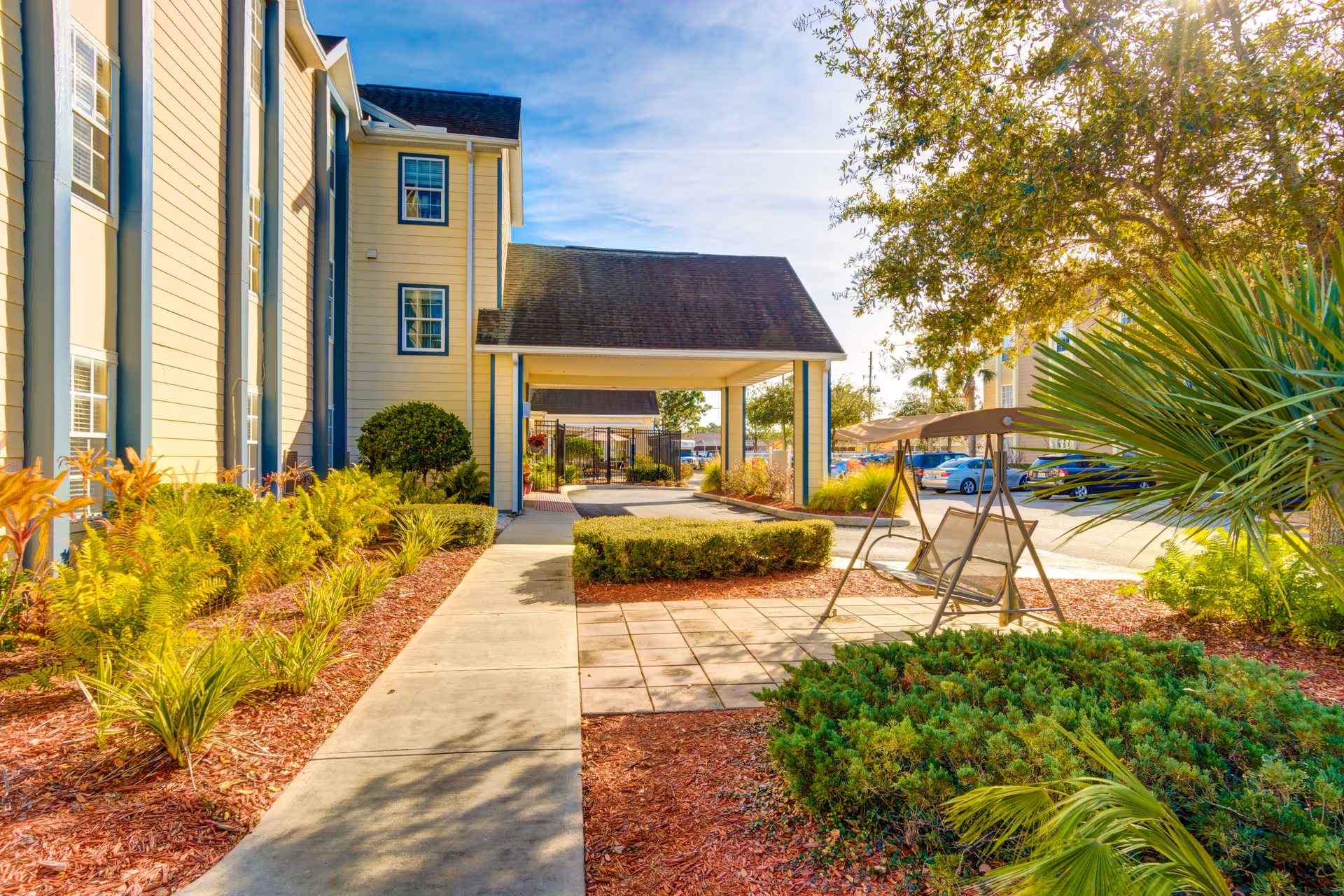 Front entrance and landscaped walkway of a senior living building with a covered entry, swinging bench and parking area.