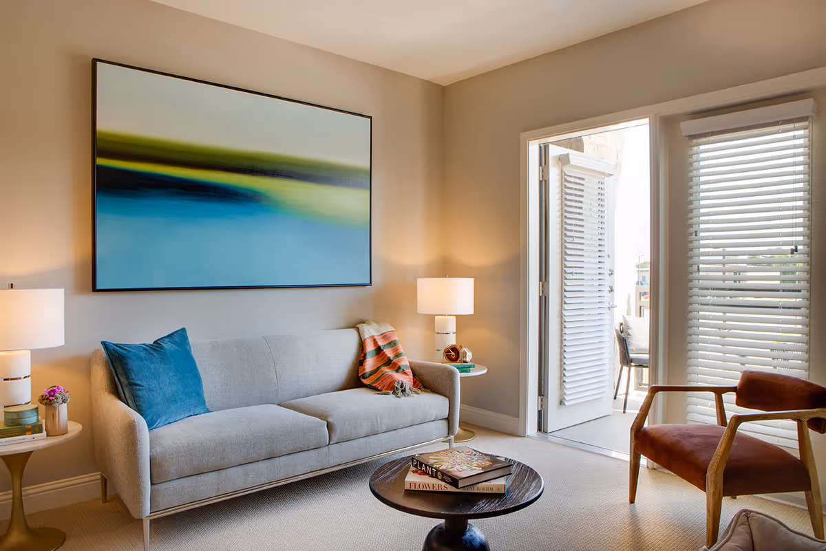 A cozy living room with a light gray sofa adorned with a blue pillow and a striped throw blanket. A round wooden coffee table in front of the sofa holds two books. There are two side tables with lamps on either side of the sofa. A large abstract painting with blue, green, and yellow hues hangs on the wall above the sofa. To the right, there is a door and window with white blinds leading to an outdoor patio with chairs.