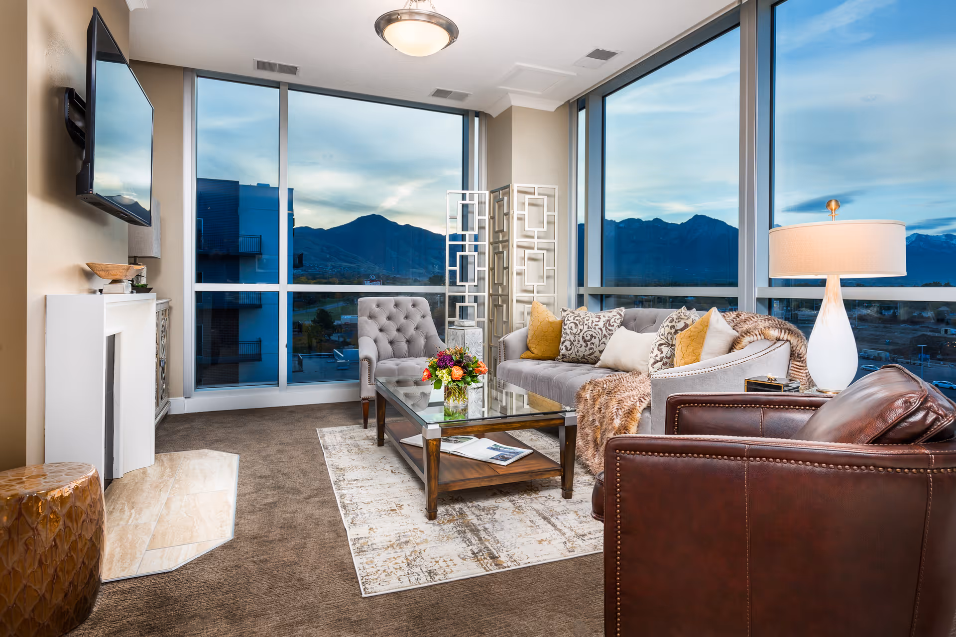 A bright and modern living room with large floor-to-ceiling windows offering a view of mountains. The room features a gray tufted armchair, a gray sofa with multiple decorative pillows and a throw blanket, a glass coffee table with a flower arrangement, a brown leather armchair, a white table lamp on a side table, a wall-mounted flat-screen TV, and a small fireplace with a decorative bowl on top.