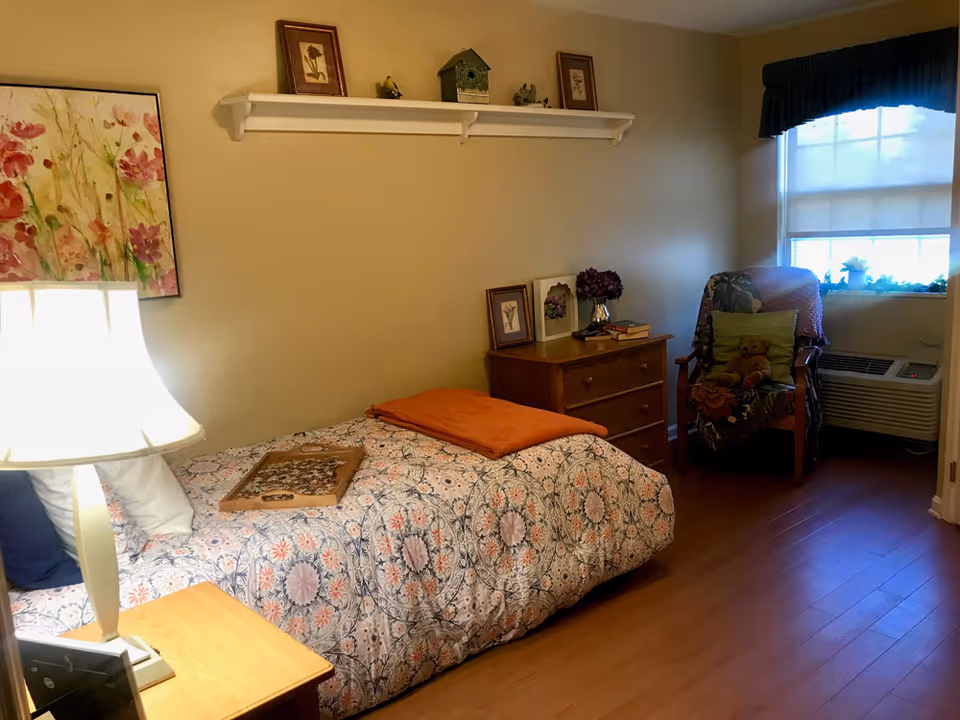 A cozy bedroom with a single bed covered in a patterned bedspread and an orange blanket folded at the foot. A wooden nightstand with a lamp is beside the bed. On the wall above the bed is a white shelf holding framed pictures and decorative items. Across from the bed is a wooden dresser with framed pictures and a vase of flowers. A cushioned armchair with a teddy bear and a blanket sits near a window with a dark valance and a white shade. The floor is wooden.
