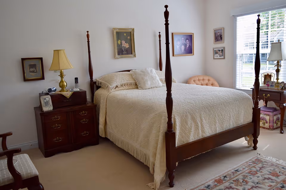 A cozy bedroom with a wooden four-poster bed covered in a cream-colored quilt and pillows. There are two wooden nightstands on either side of the bed, each with a lamp and framed photos. The room has light-colored walls adorned with several framed pictures and a window with white blinds letting in natural light. A patterned rug is partially visible on the carpeted floor.