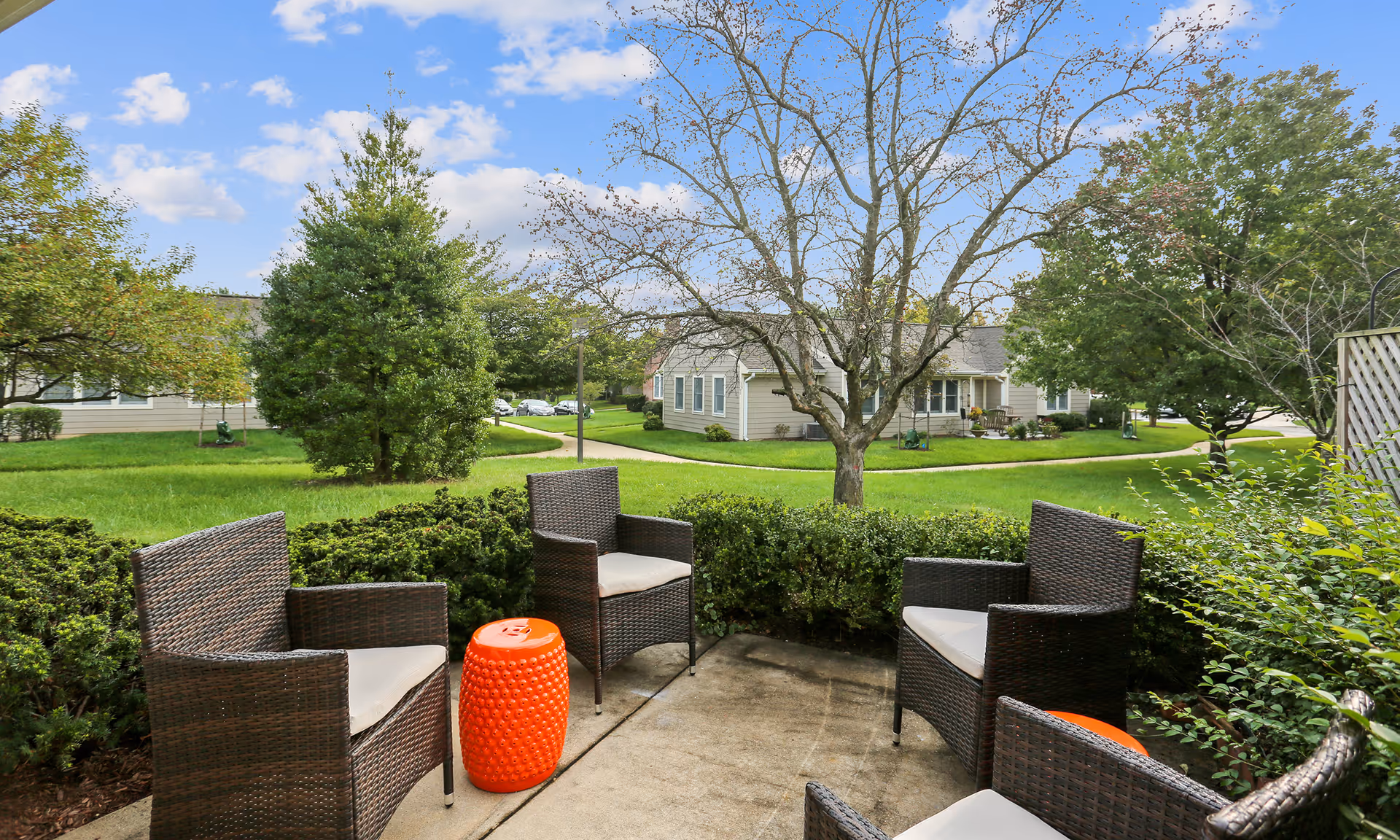 Patio with wicker chairs and orange stools facing a grassy courtyard and single-story residence buildings.