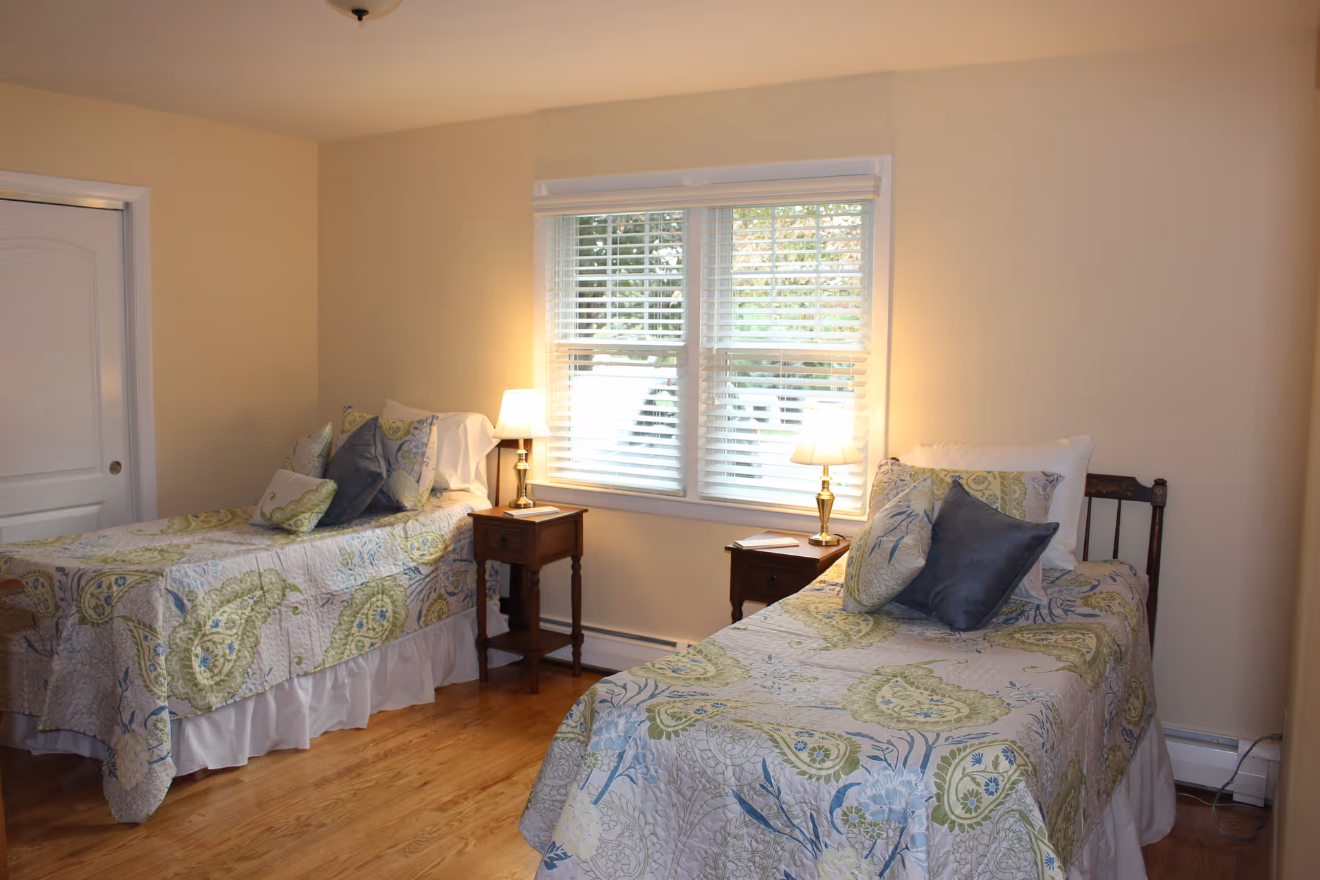 A bedroom with two twin beds, each with patterned bedspreads and multiple pillows. Between the beds are two wooden nightstands, each with a lamp. A window with white blinds is centered on the wall behind the nightstands, letting in natural light. The room has light-colored walls and wooden flooring.