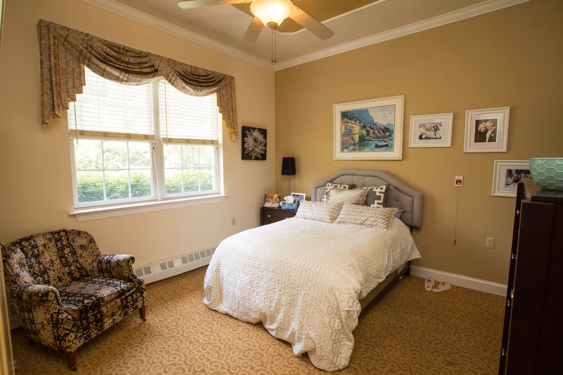 A cozy bedroom with a large window covered by patterned valances, a comfortable bed with white bedding and multiple pillows, a patterned armchair, a nightstand with framed photos, a ceiling fan with a light, and several framed pictures on the beige walls.