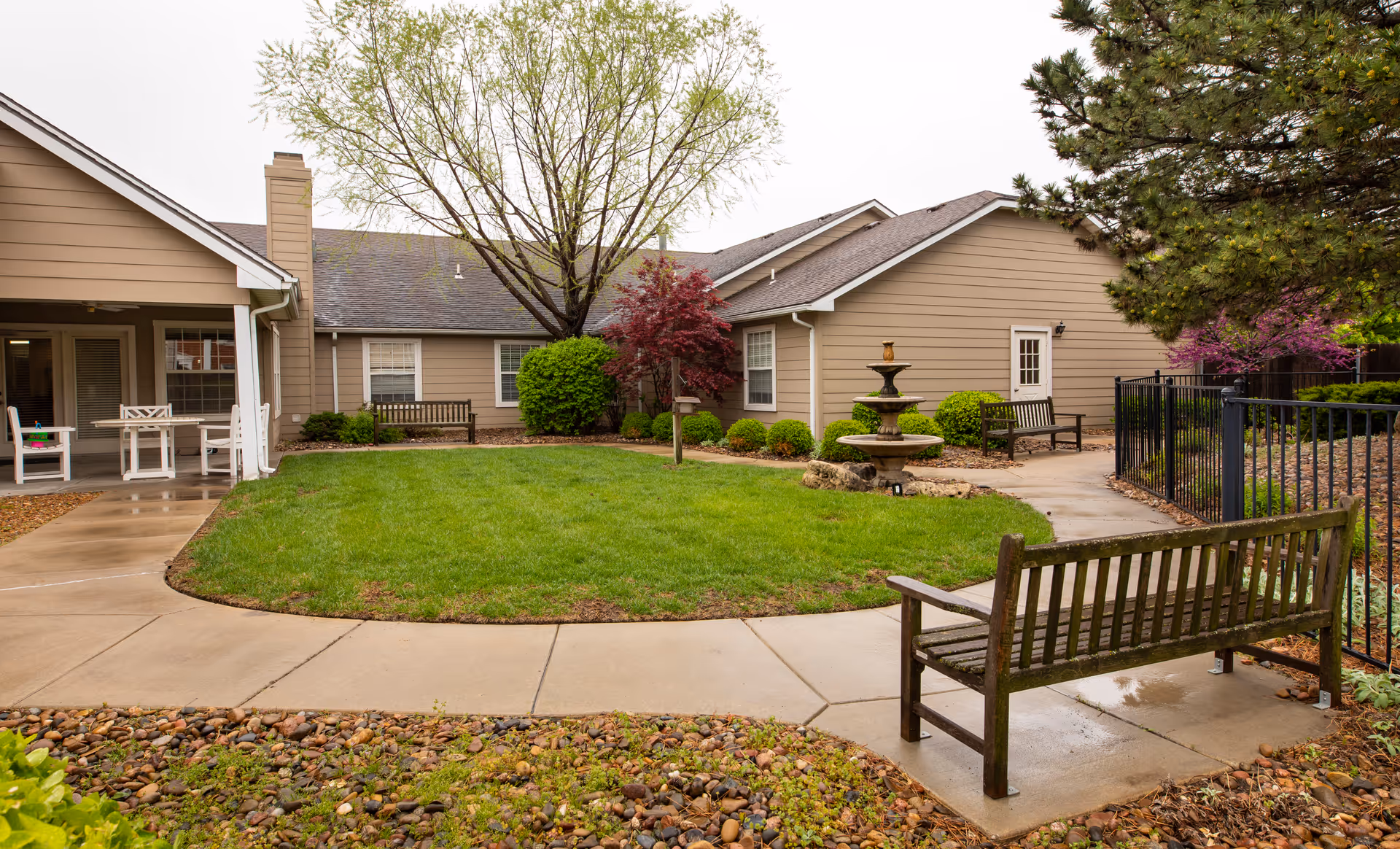 Outdoor courtyard area of Homestead Assisted Living & Memory Care of Augusta featuring a green lawn surrounded by a concrete walkway, wooden benches, a multi-tiered stone fountain, and various trees and shrubs. The building exterior is beige with white trim, and there is a covered patio area with white chairs and tables on the left side.