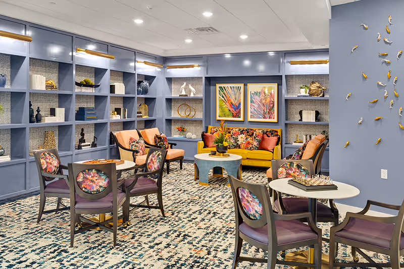 Bright, colorful communal lounge with patterned chairs, a yellow sofa, built-in blue bookshelves, and decorative wall art.