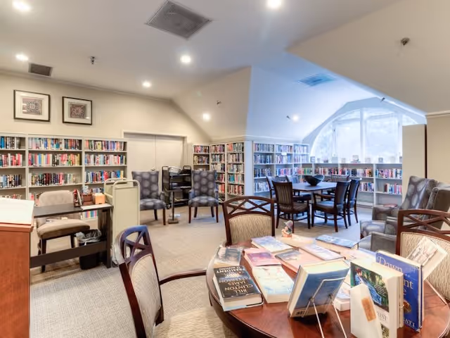 A bright and cozy library room with bookshelves lining the walls filled with books. Several chairs and tables are arranged for reading and socializing, including a round table in the foreground with books displayed on it. Large arched windows allow natural light to fill the space.