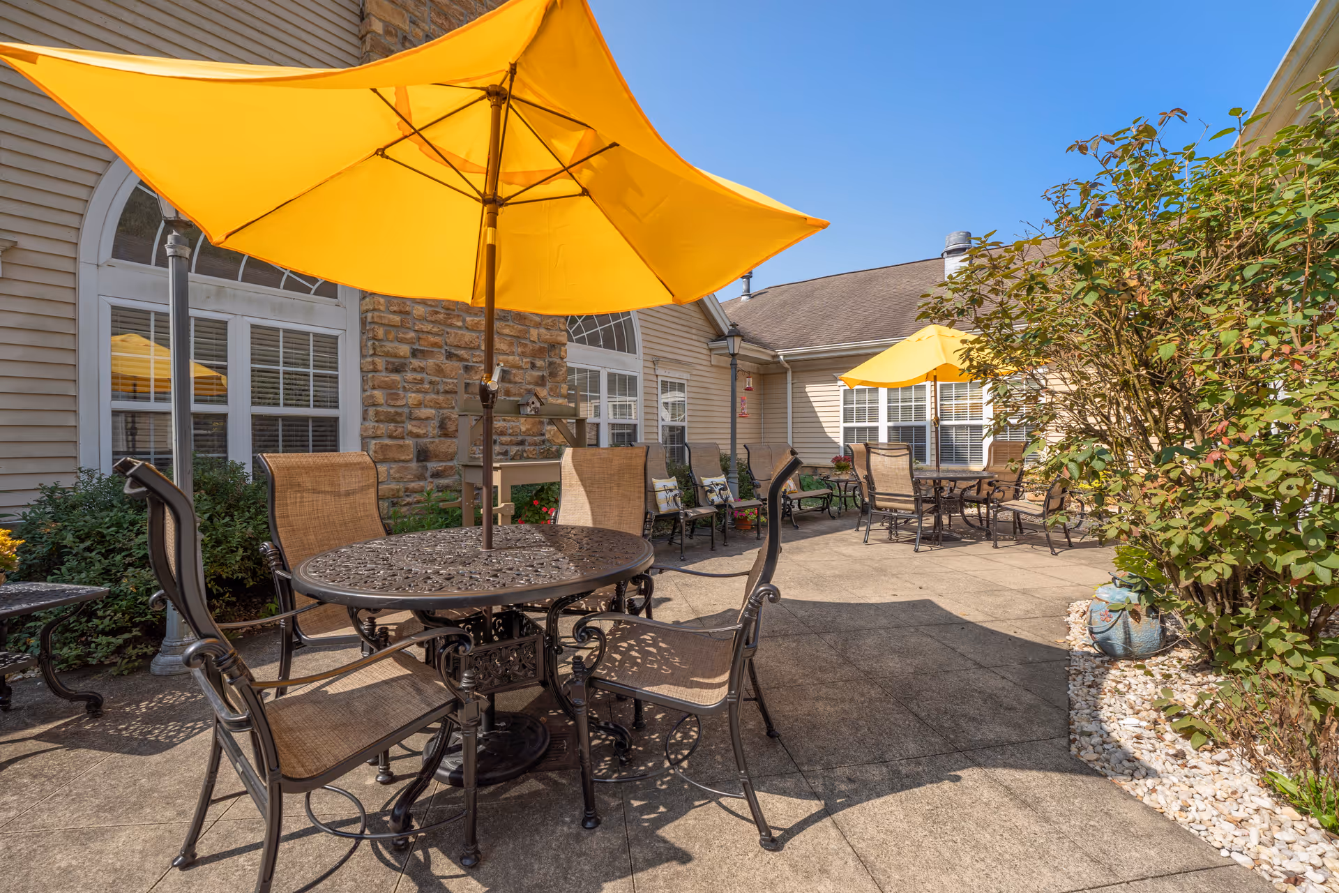 Outdoor patio area with metal tables and chairs, each table shaded by a large yellow umbrella. The patio is adjacent to a building with beige siding and stone accents, surrounded by greenery and plants under a clear blue sky.