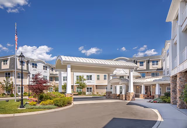 Exterior view of a senior living facility with a covered entrance driveway, landscaped garden areas, and a clear blue sky with some clouds.