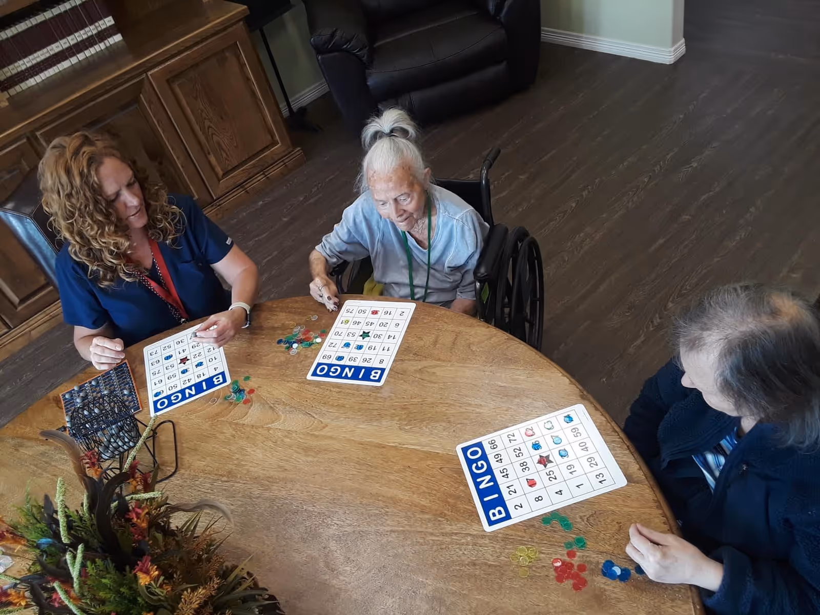 Three women sitting around a wooden round table playing bingo. One woman is in a wheelchair, and another appears to be a caregiver. Bingo cards and colorful markers are on the table. The room has wooden flooring and wooden furniture in the background.