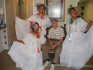An elderly man sitting on a chair indoors, surrounded by three women dressed in traditional white dresses with colorful floral headpieces and jewelry, posing and smiling.
