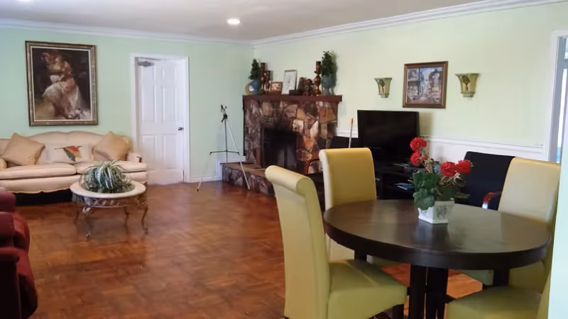 A cozy living room with a round wooden table surrounded by four yellow chairs in the foreground. A potted plant with red flowers is on the table. In the background, there is a beige sofa with cushions, a stone fireplace with decorative items on the mantel, a flat-screen TV on a stand, and framed artwork on the walls. The room has wooden flooring and light green walls.
