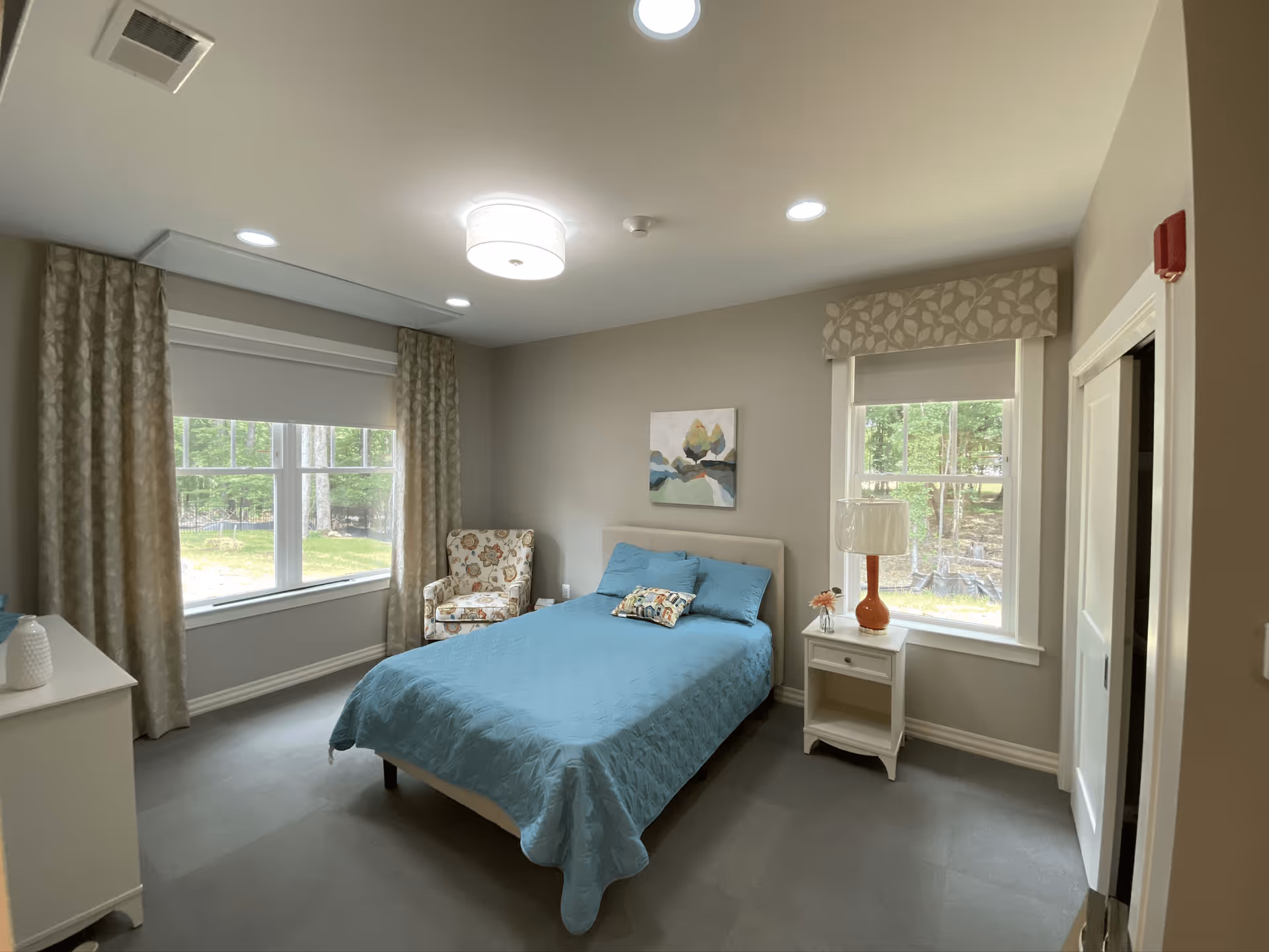 Bright bedroom with a blue-covered bed, patterned armchair, nightstand with lamp, and two large windows showing trees outside.