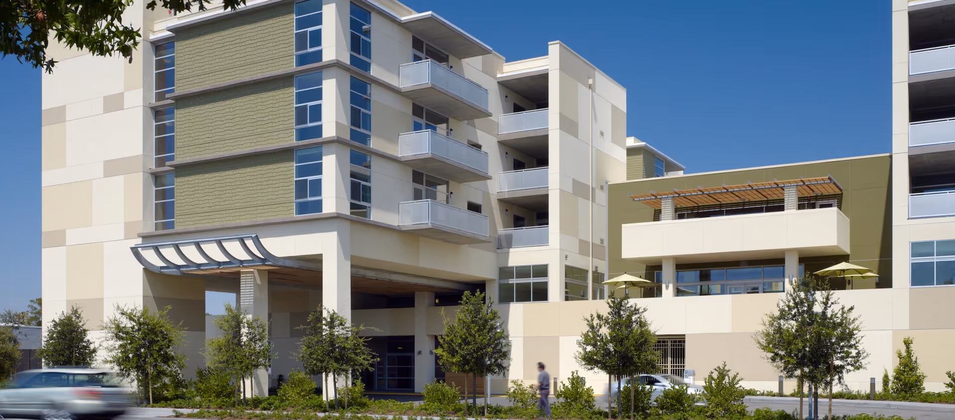 Exterior view of a modern multi-story senior living facility building with balconies, large windows, and a covered entrance. There are small trees and landscaping in front, a person walking on the sidewalk, and a car driving by on the street.