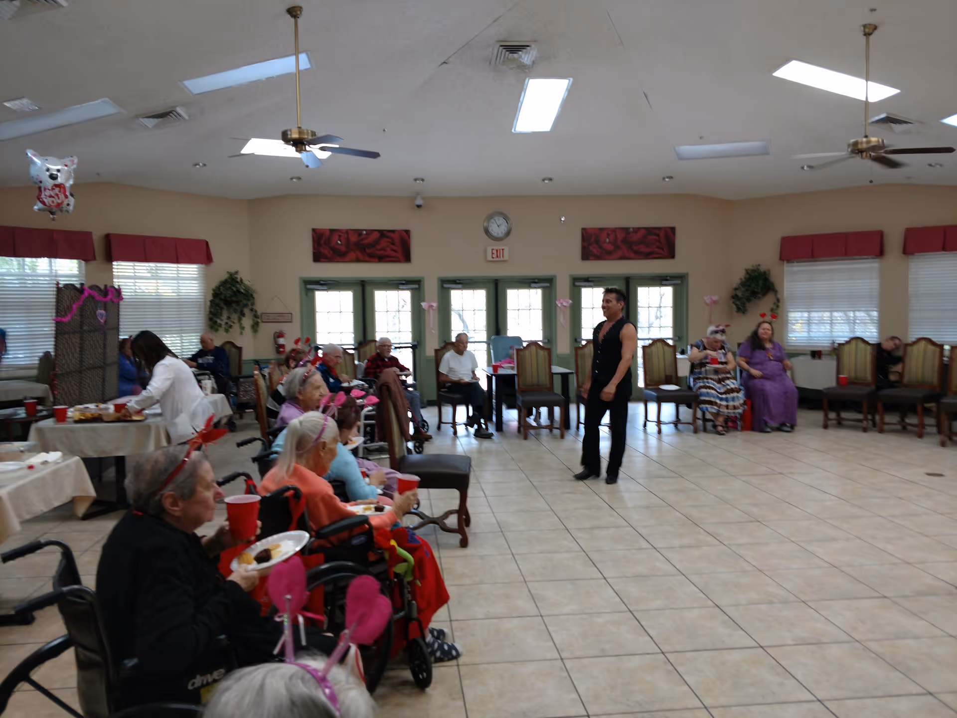 A group of elderly people seated in a spacious, well-lit room with tiled floors and ceiling fans. Some are in wheelchairs, holding plates and red cups, while a man stands in the center of the room. The room has large windows with red valances and green doors at the back. Decorations suggest a festive or social event.