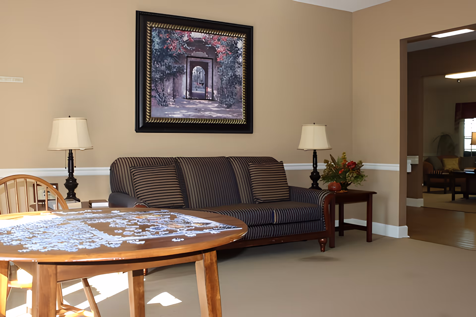 A cozy living room area with a striped sofa, two table lamps on either side, a wooden table with a lace tablecloth, and a framed painting of an archway with red flowers on the wall. The room has beige walls and carpeted floor, with an open doorway leading to another room with additional seating.