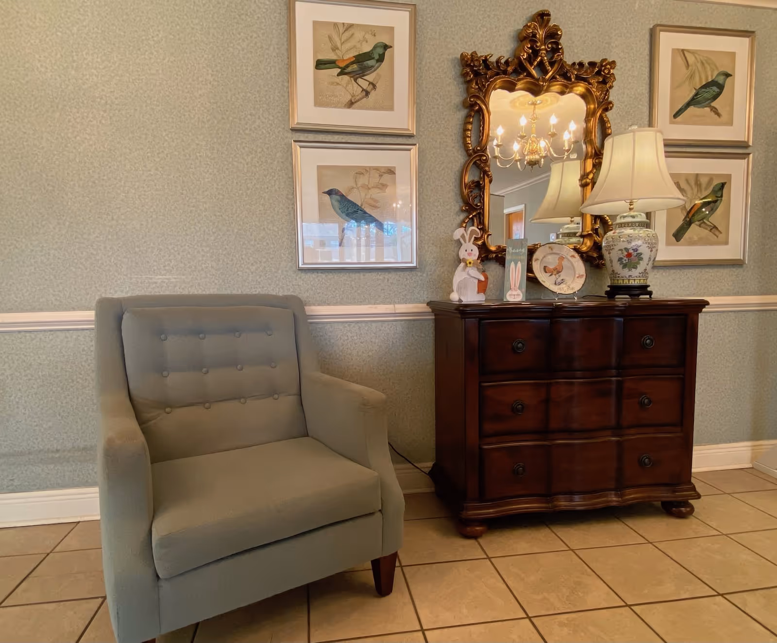 A cozy interior corner featuring a light blue upholstered armchair next to a dark wooden chest of drawers. On top of the chest is a decorative table lamp with a floral design, a gold ornate mirror, a decorative plate, and two bunny figurines. The wall behind has a light blue patterned wallpaper with four framed bird illustrations arranged around the mirror. The floor is tiled.