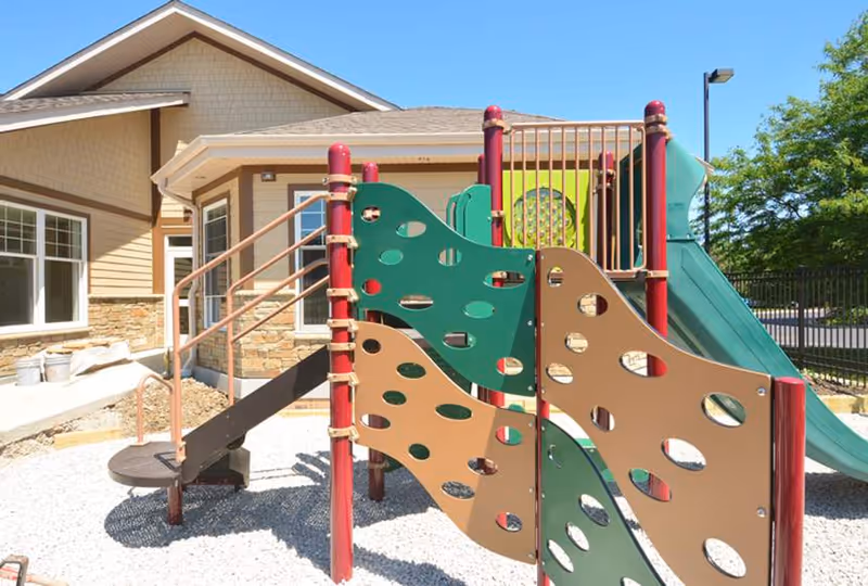 Outdoor playground area with colorful climbing structures and slides in front of a beige building with stone accents under a clear blue sky.