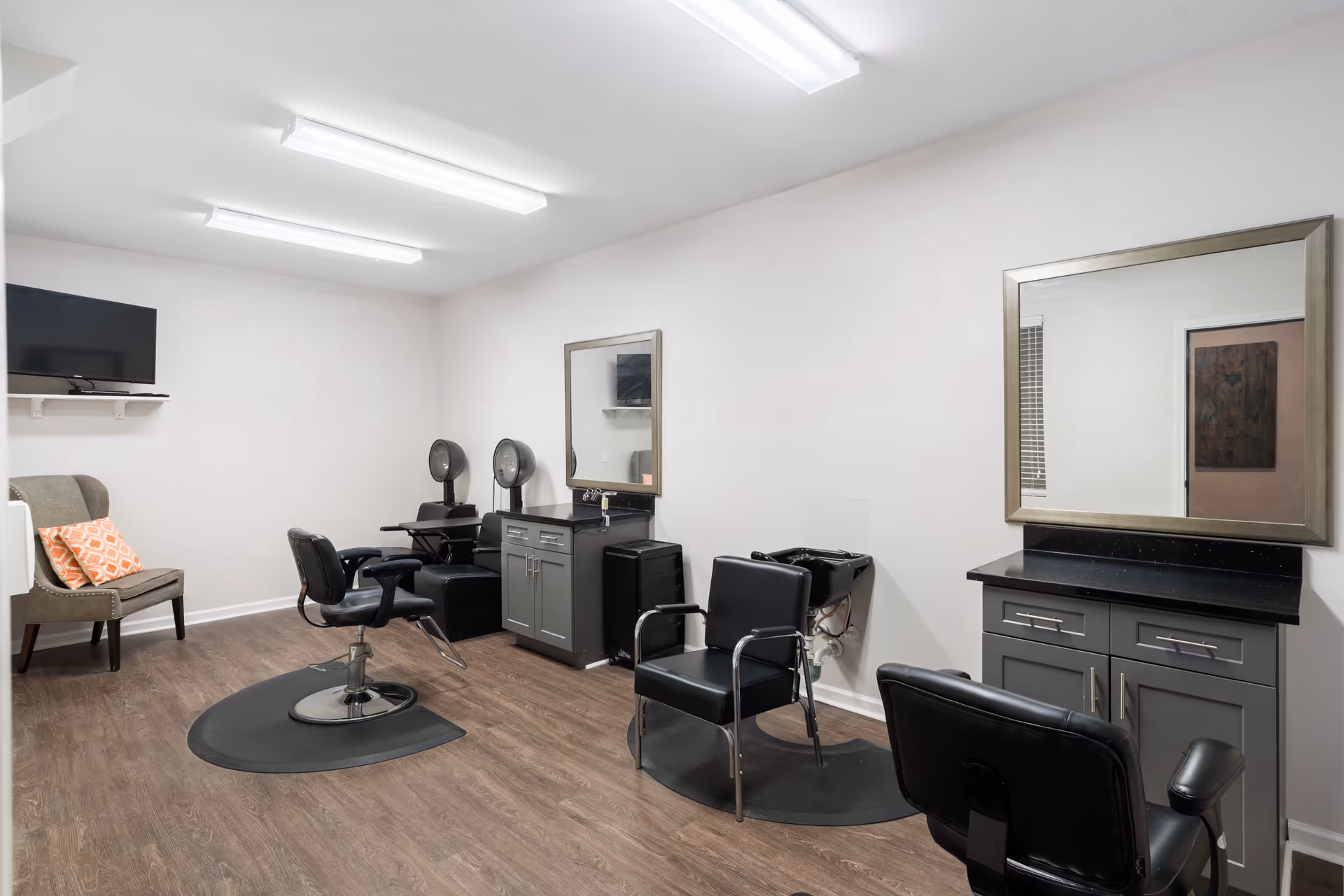 A hair salon room with styling chairs, mirrors, hooded dryers and a shampoo sink in a senior living facility.
