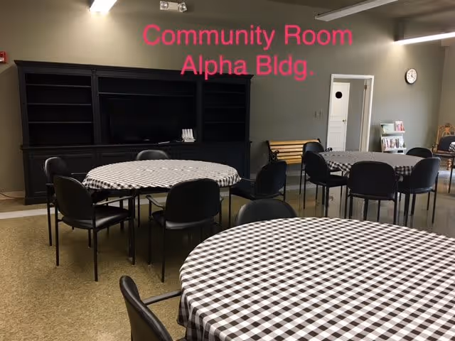 Interior view of a community room with round tables covered in black and white checkered tablecloths, surrounded by black chairs. There is a large black shelving unit against the wall, a bench, a clock, and a magazine rack. The wall has text that reads 'Community Room Alpha Bldg.'