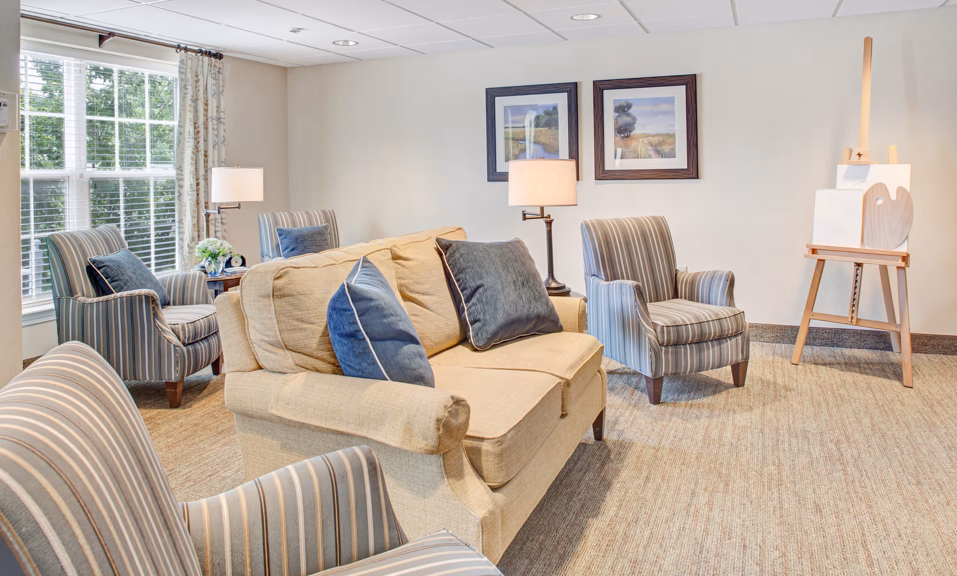 A cozy living room with a beige sofa adorned with blue and gray pillows, surrounded by four striped armchairs. Two framed landscape paintings hang on the wall above a floor lamp. A wooden easel with a blank canvas and a heart-shaped palette is positioned in the corner. Large windows with white blinds and patterned curtains allow natural light to fill the room.