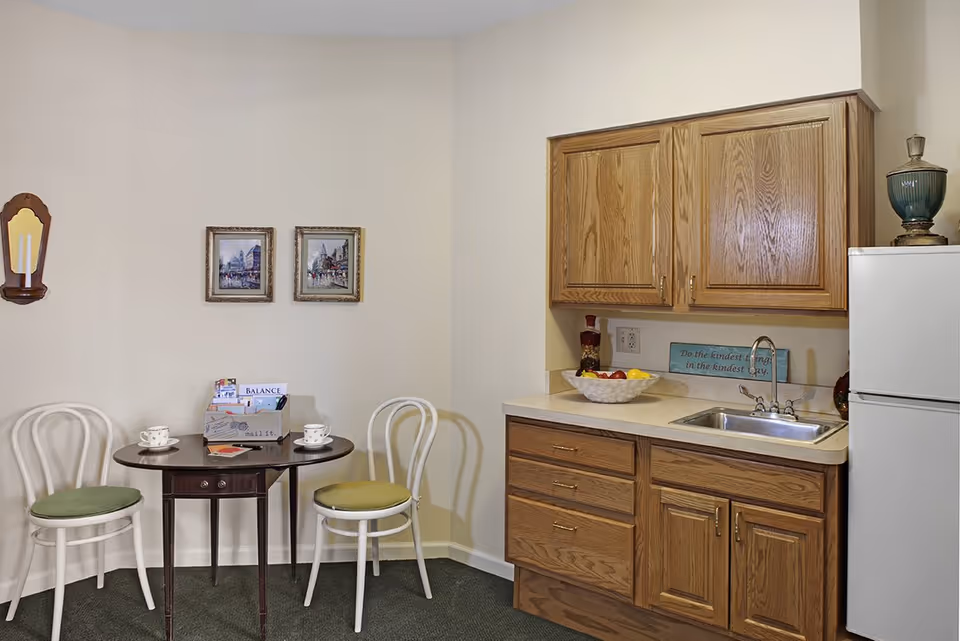 Small kitchenette and dining nook with oak cabinets, a sink and refrigerator on the right and a small table with two chairs and wall art on the left.