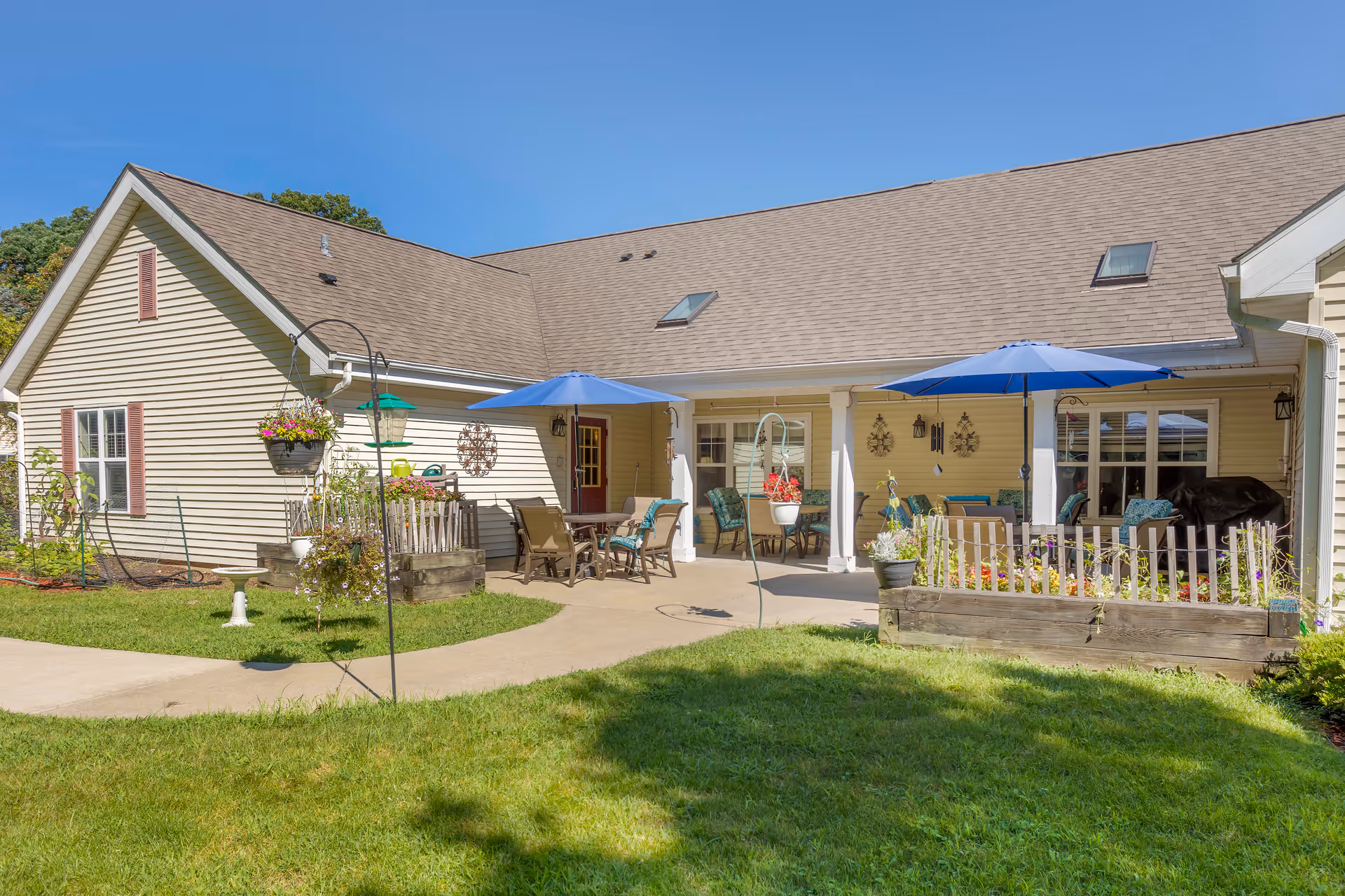 Outdoor patio area of a senior living facility with beige siding building, two blue umbrellas shading tables and chairs, flower beds, hanging plants, and a well-maintained lawn under a clear blue sky.