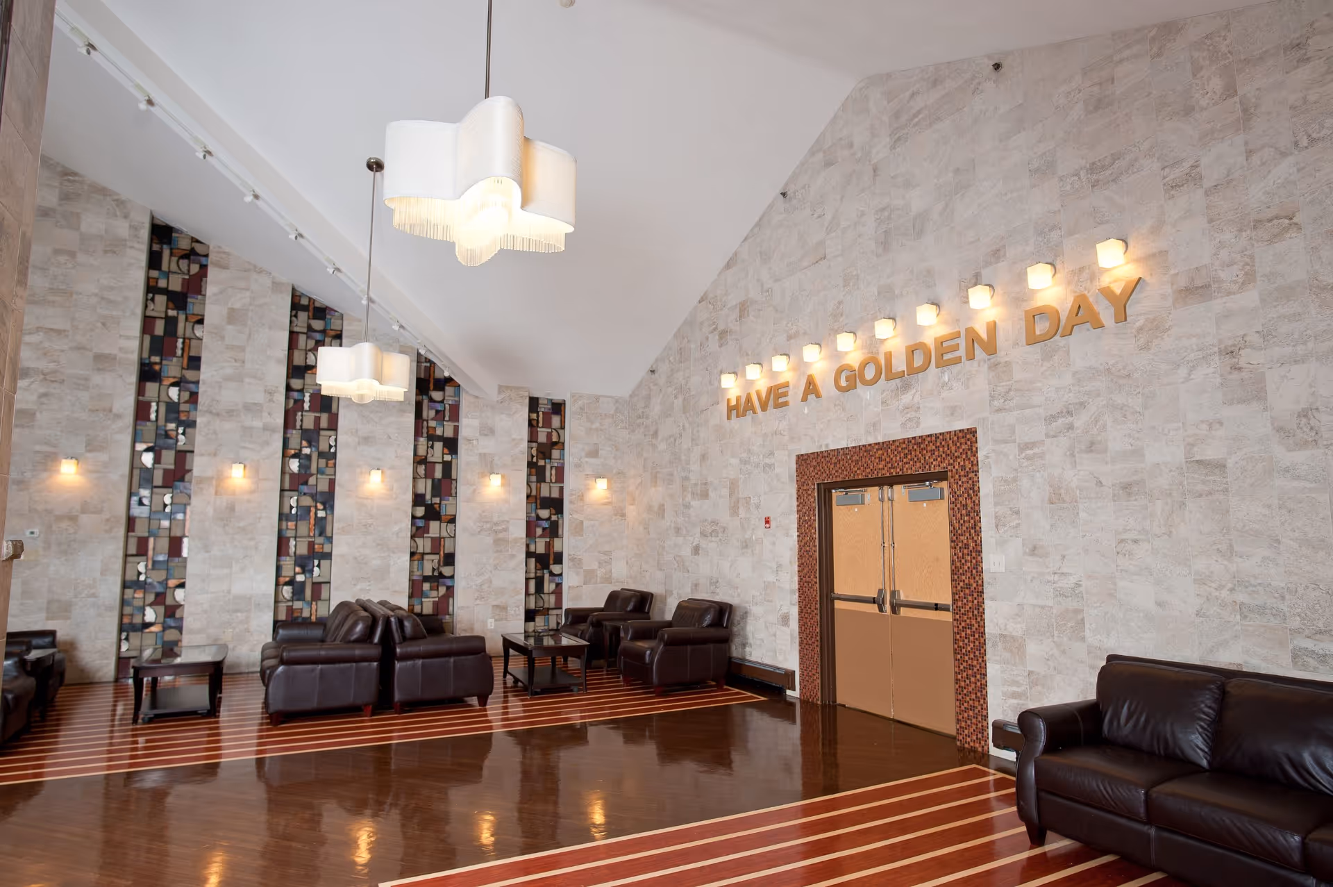 A spacious lounge area with high ceilings, featuring dark leather sofas and armchairs arranged around small tables. The walls are decorated with vertical stained glass panels and light fixtures. Above a double door, illuminated letters spell out 'HAVE A GOLDEN DAY'. The floor has a polished wood finish with light-colored stripes.