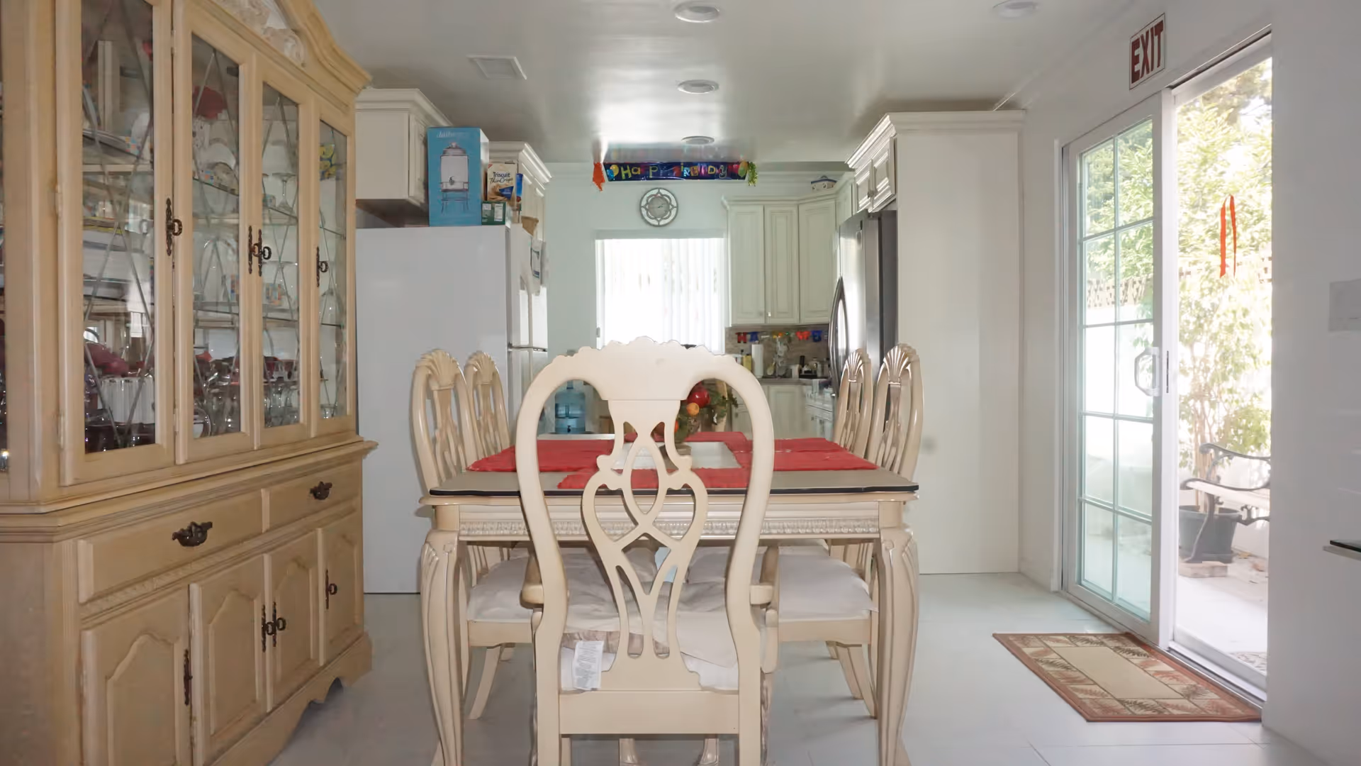 A dining area with a wooden dining table and six matching chairs with white cushions. The table has red placemats and a centerpiece. To the left is a wooden china cabinet with glass doors displaying glassware. In the background, there is a kitchen with white cabinets, a refrigerator, and a window with vertical blinds. On the right side, there is a sliding glass door leading to an outdoor area with plants and a bench. An exit sign is visible above the sliding door.