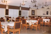 Dining room with multiple tables set with white tablecloths, wooden chairs, chandeliers, and windows with blinds.