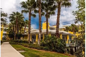 Yellow multi-story building with palm trees and a sidewalk in front under a cloudy sky.
