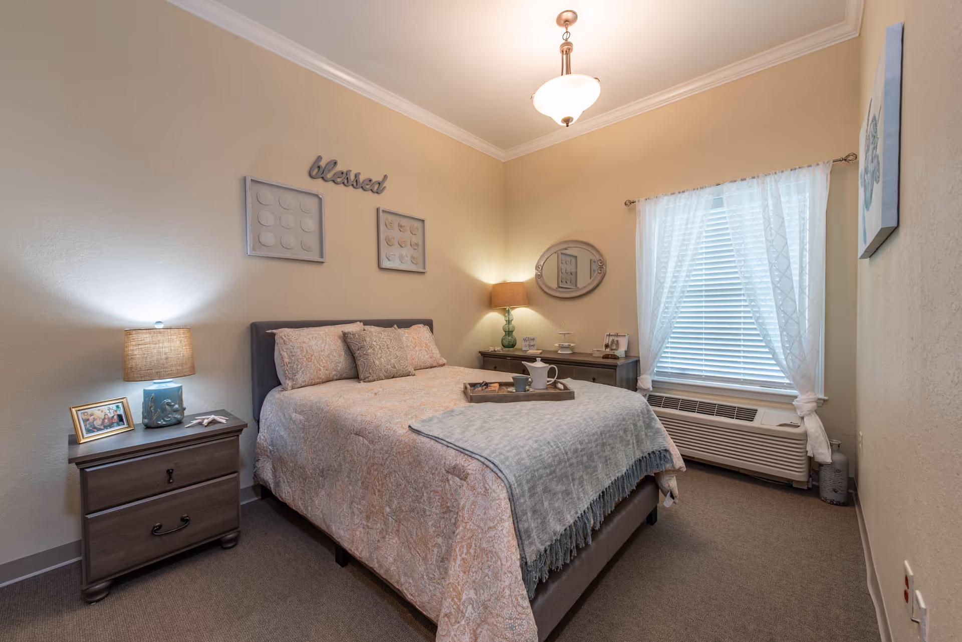 A cozy furnished bedroom featuring a made bed with pillows, two nightstands with lamps, a window with blinds and wall decor that reads 'blessed'.