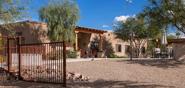 Exterior view of a single-story senior living facility building with a gated entrance, desert landscaping including trees and shrubs, and outdoor seating under an umbrella on a sunny day with a blue sky and scattered clouds.