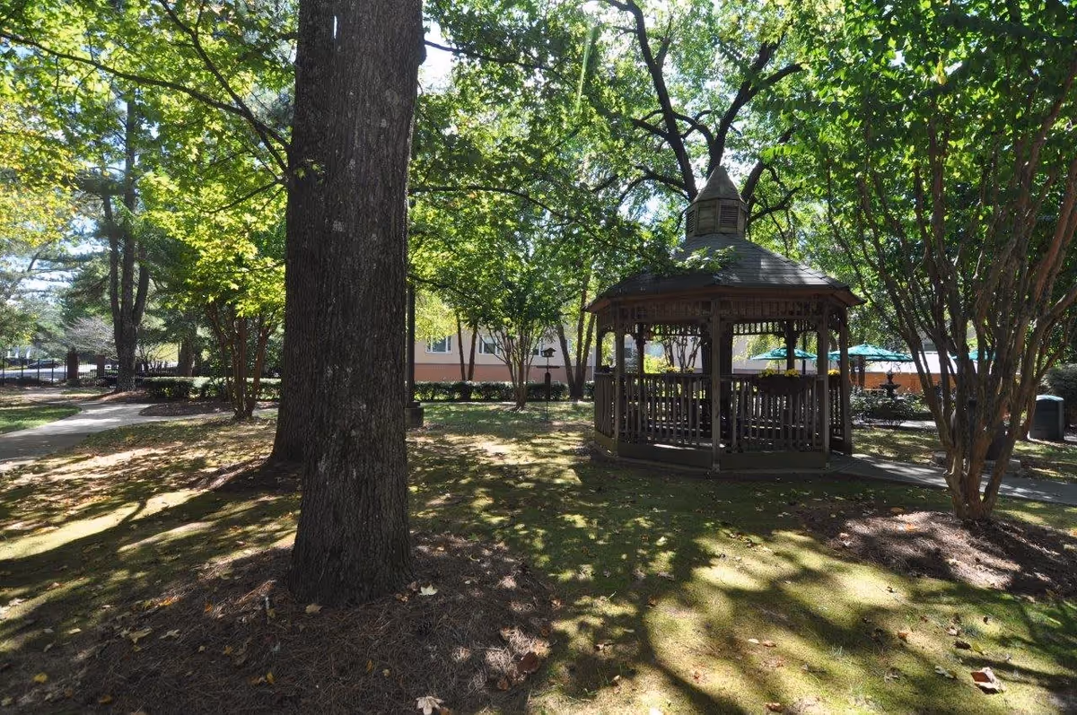 A wooden gazebo in a shaded garden surrounded by trees with a walkway and a building in the background.