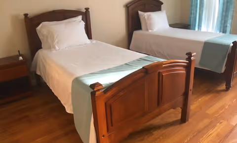 A bedroom with two single wooden beds, each made with white sheets, pillows, and light blue bed runners. There is a wooden nightstand between the beds and a window with light blue curtains on the right side.