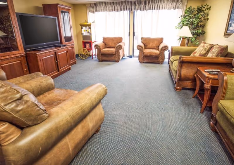 A cozy living room with two brown leather armchairs, a matching leather sofa, a wooden entertainment center with a large flat-screen TV, a side table with a lamp, a potted plant, and a popcorn machine near a large window with curtains.