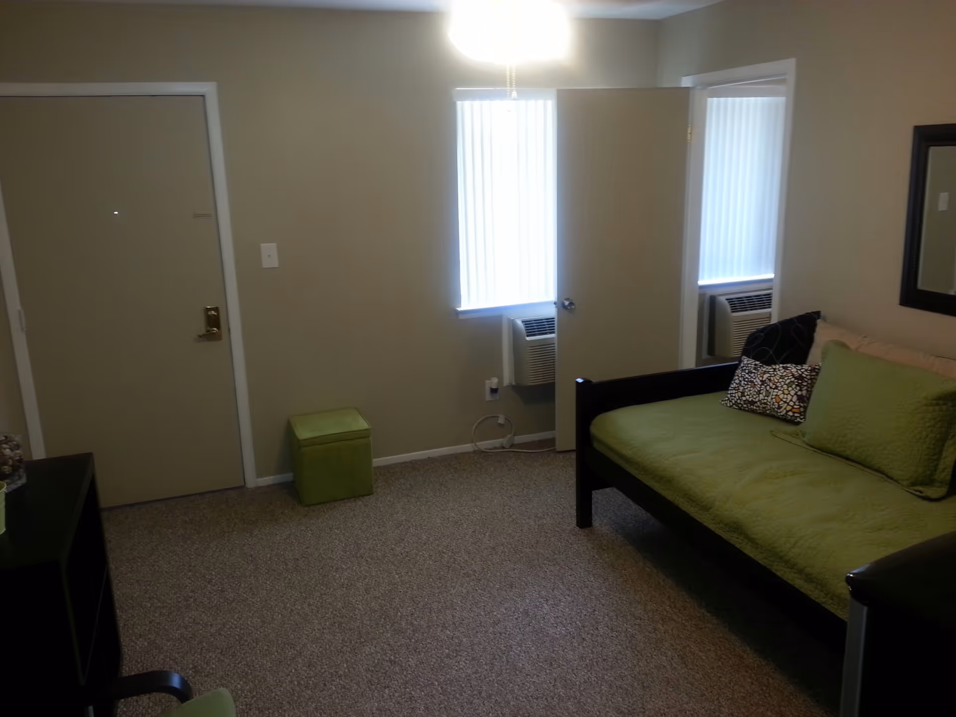 Small studio living room with a green daybed, entrance door, two narrow windows with vertical blinds and wall air conditioners.