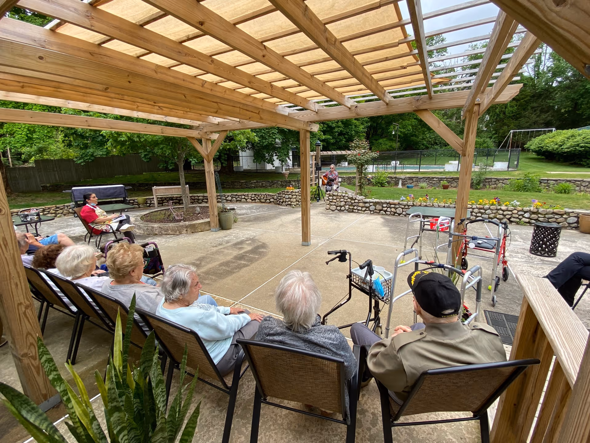 Seniors seated under a wooden pergola on a patio, facing a performer in the outdoor courtyard.