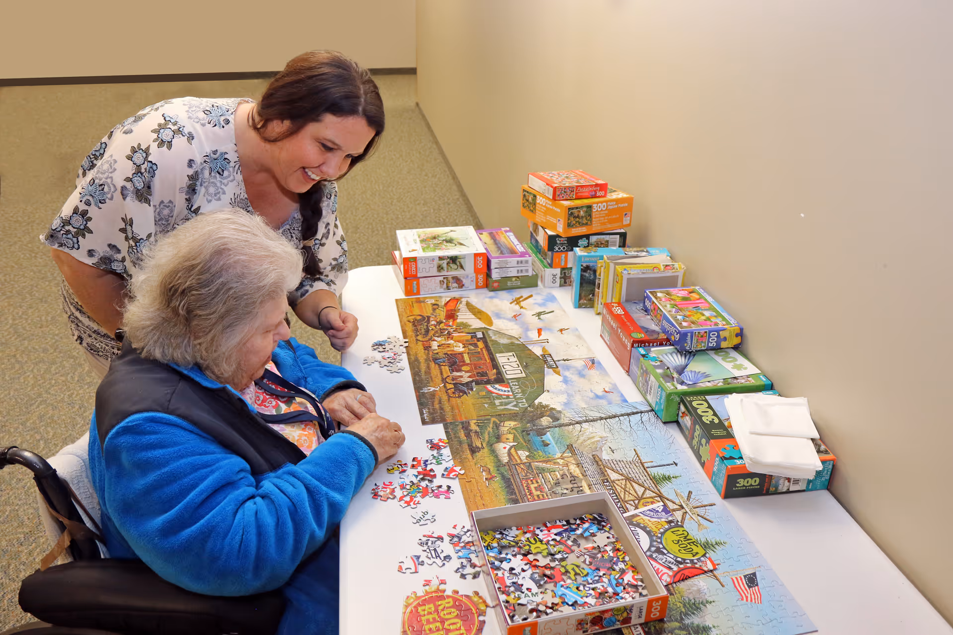 An elderly woman in a wheelchair and a younger woman are working together on assembling a large jigsaw puzzle on a table. The table is also stacked with several other puzzle boxes. They are in a carpeted indoor space with beige walls.