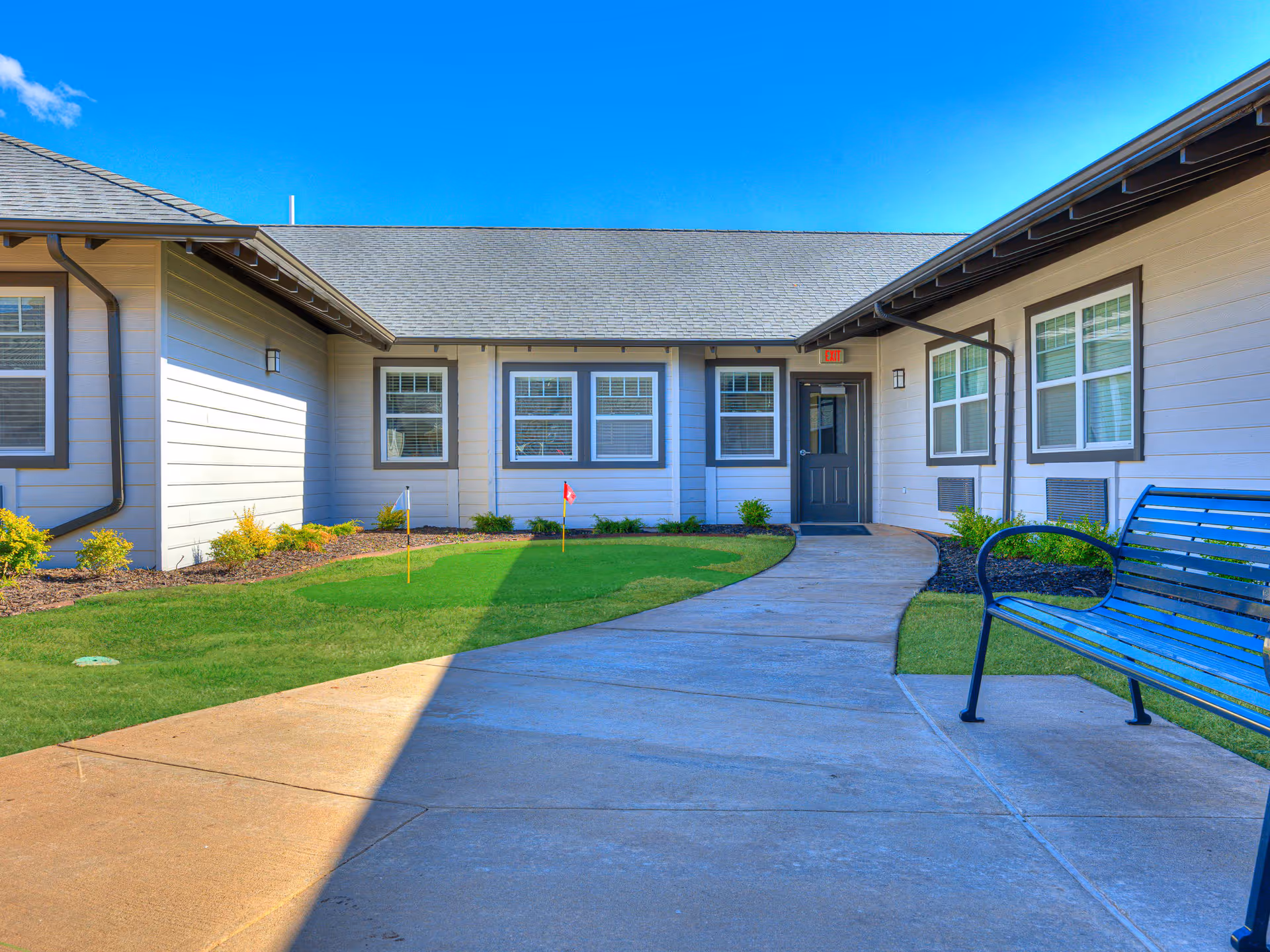 Outdoor courtyard area of a senior living facility with a concrete walkway, green grass, small putting green with flags, a blue bench, and light-colored building walls with multiple windows under a clear blue sky.