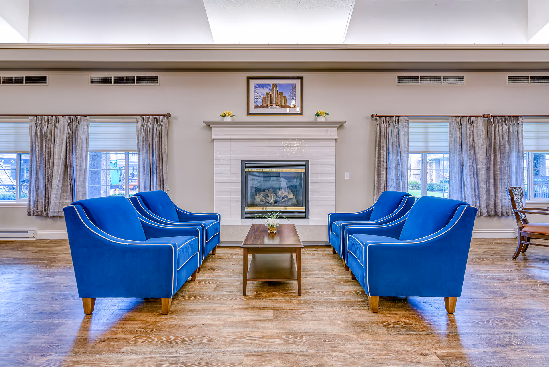 A bright common lounge with four blue armchairs arranged around a wooden coffee table facing a central fireplace.