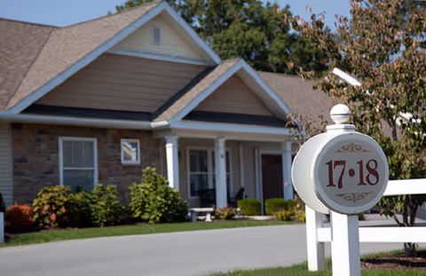 Exterior view of a residential-style building with a stone and siding facade, a covered porch with white columns, and a signpost in the foreground displaying the numbers 17 and 18. The area is landscaped with bushes and trees under a clear sky.