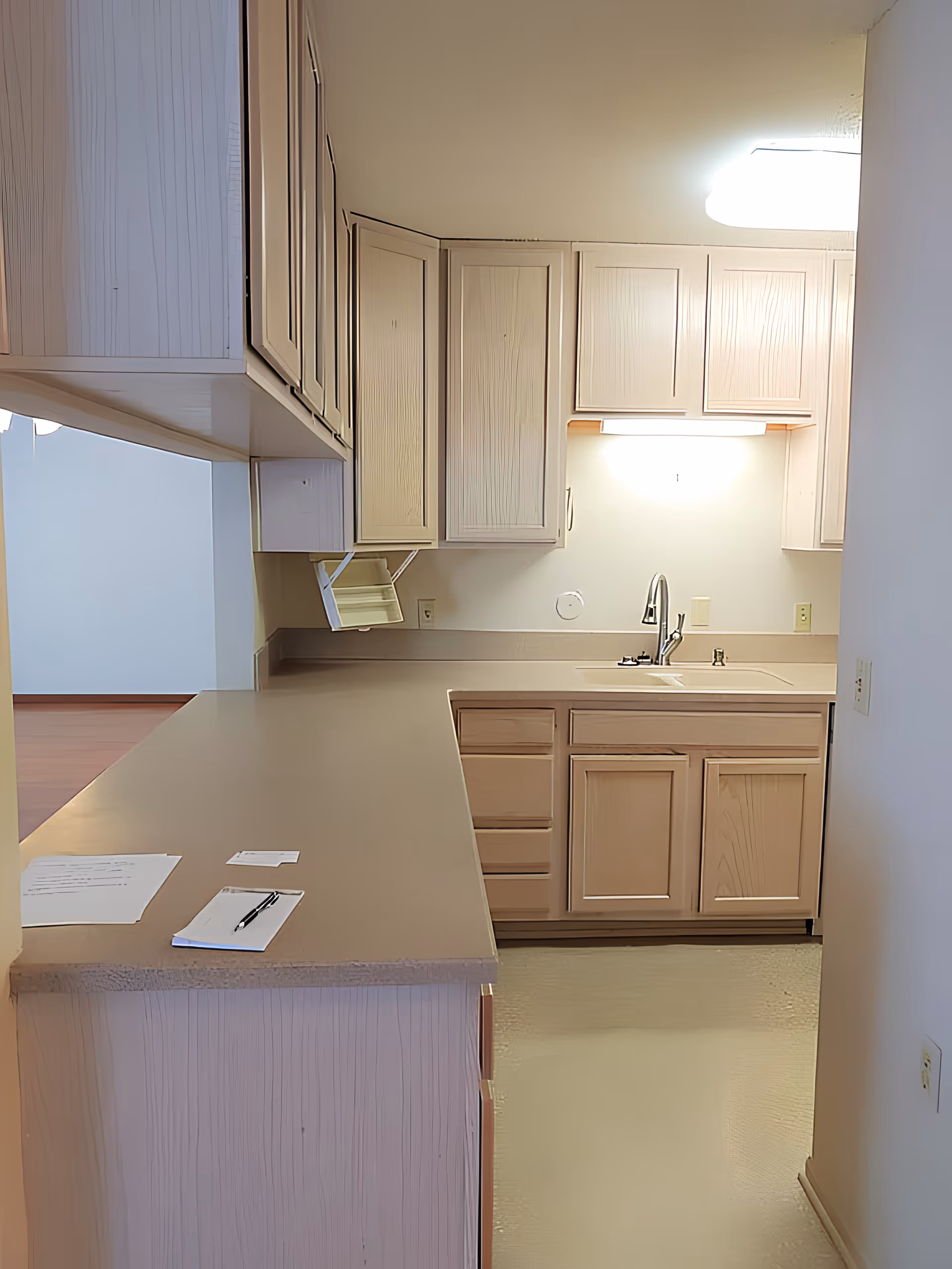 Interior view of a kitchen in Regency Retirement Residence featuring light wood cabinets, a countertop with papers and a pen on it, a sink with a faucet, and under-cabinet lighting.
