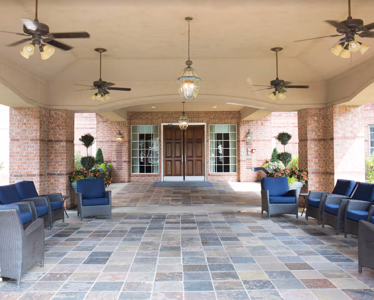 Covered building entrance with tiled floor, wicker seating with blue cushions, brick columns, ceiling fans and double wooden doors.