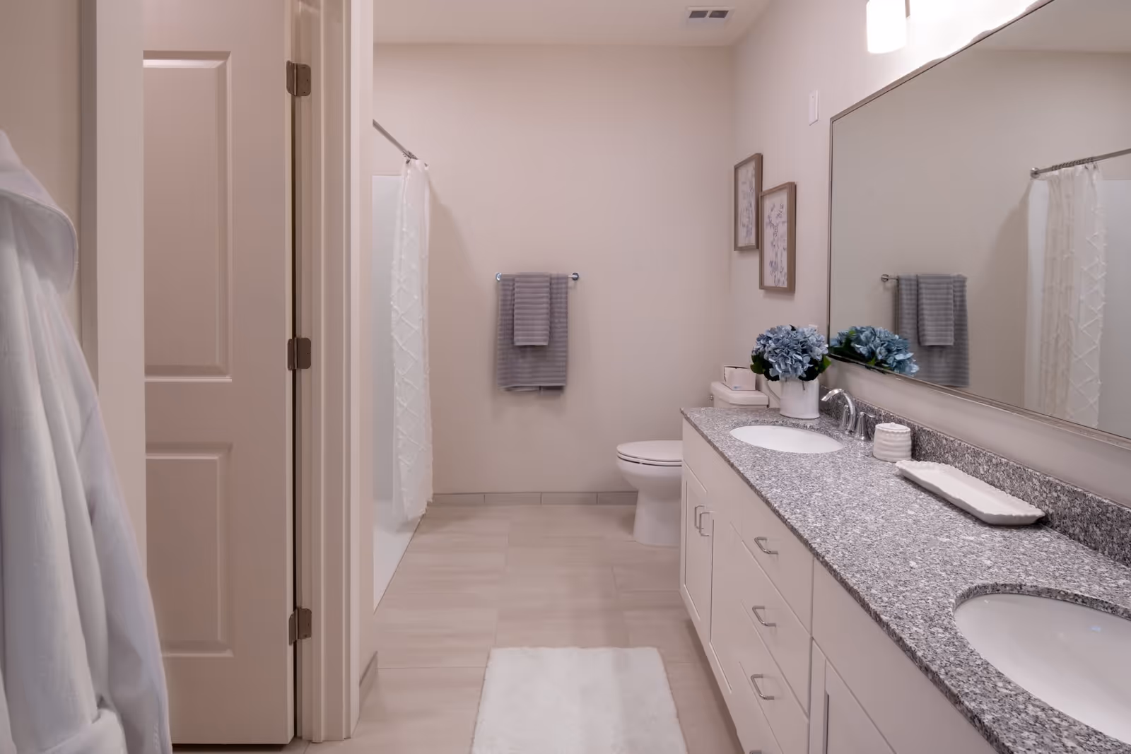 Bright modern bathroom with a double-sink granite vanity, large mirror, toilet, and shower with a white curtain.