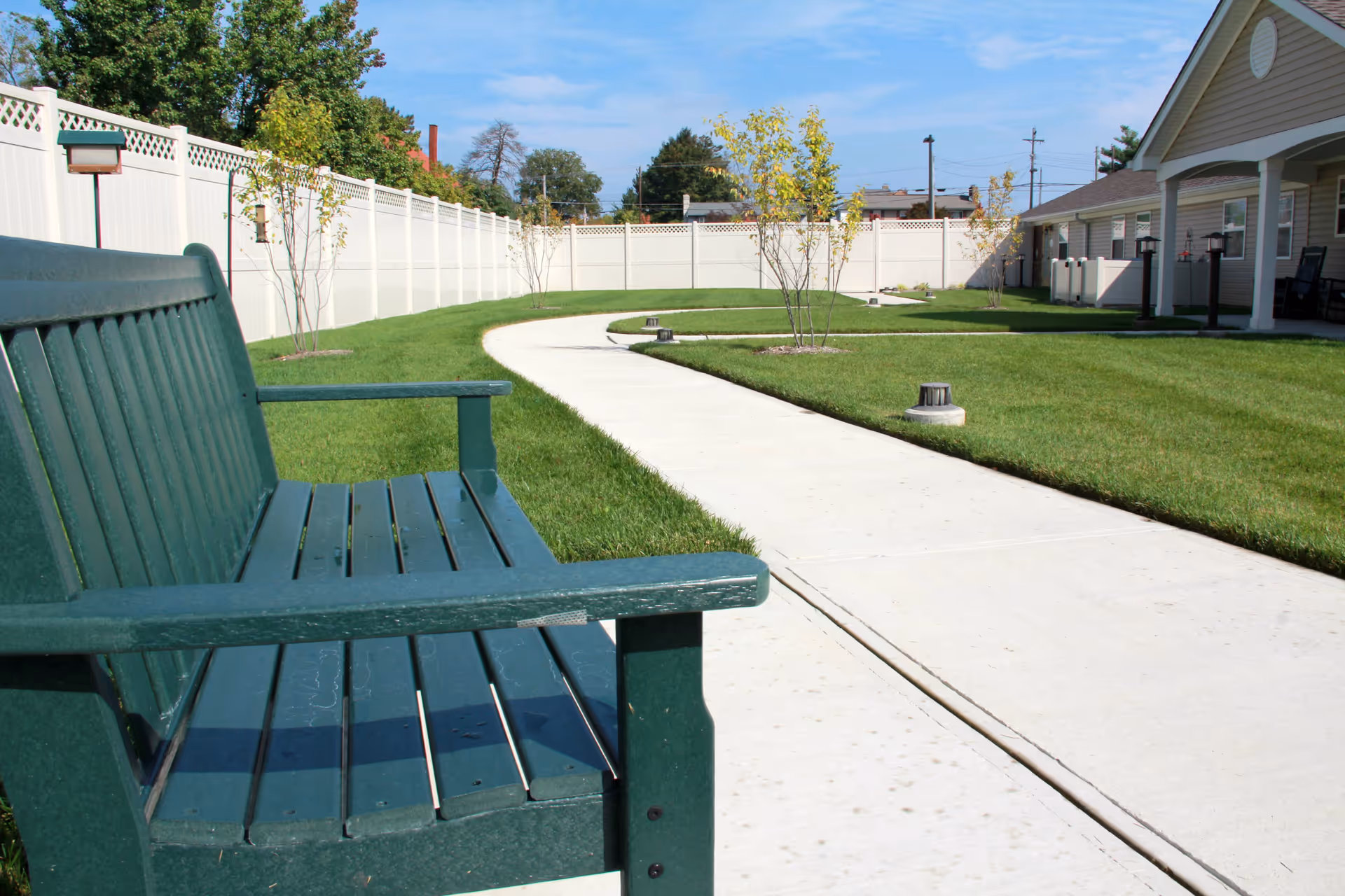 A green wooden bench next to a curved concrete pathway in a well-maintained grassy outdoor area with small trees and a white fence. A building with a covered porch is visible on the right side under a clear blue sky.