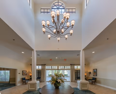 Spacious and well-lit interior lobby area with a large decorative chandelier hanging from a high ceiling. A round table with a floral arrangement is centered in the foreground. There are seating areas with chairs and lamps on either side of the room, and large windows and glass doors at the far end letting in natural light.