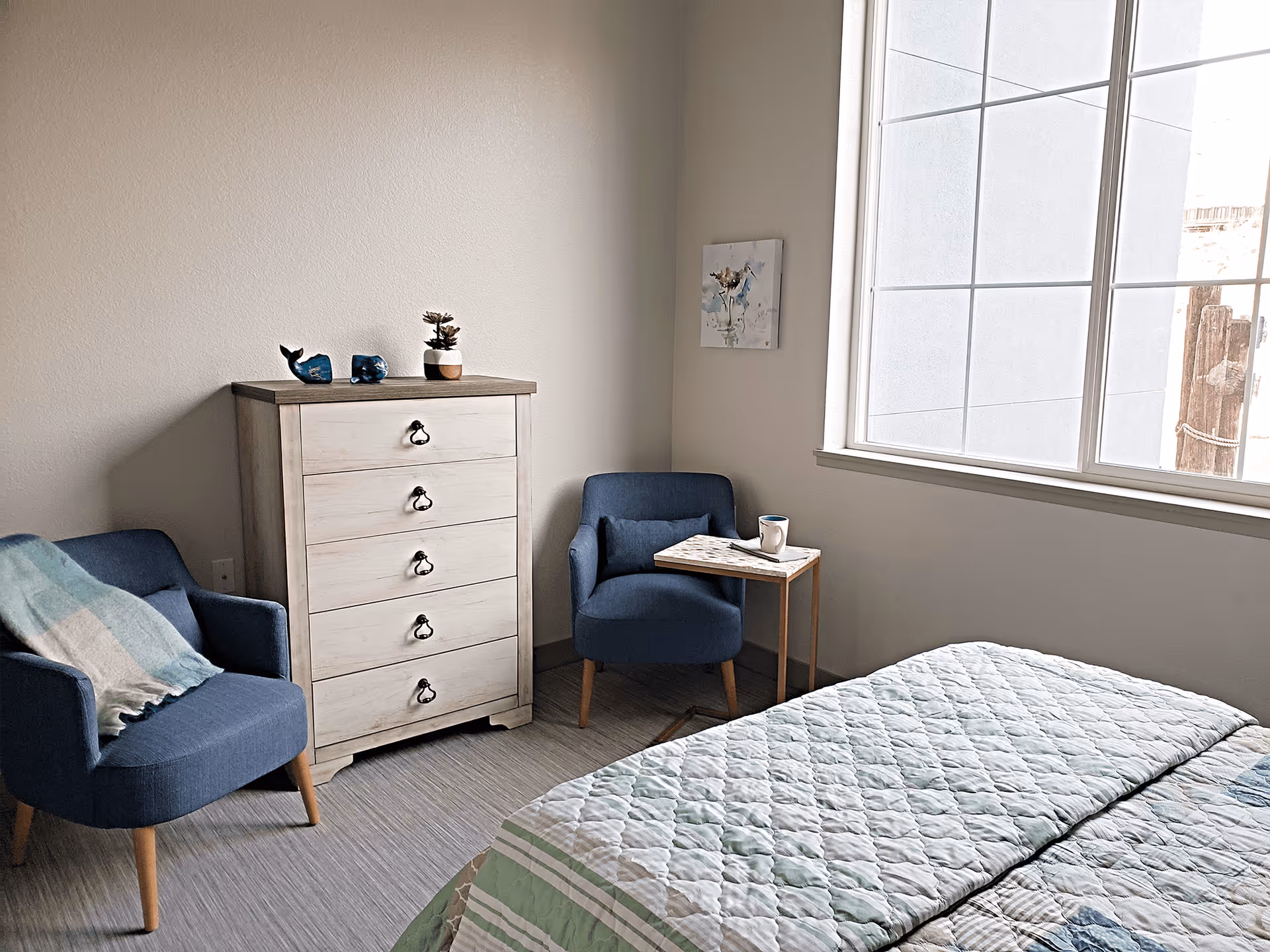 Sunlit bedroom with a bed, two blue armchairs, a wooden chest of drawers and a large window.